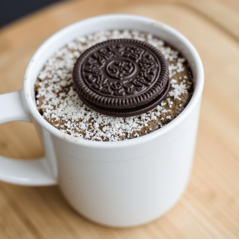 A freshly made Oreo mug cake in a white ceramic mug, topped with whipped cream, crushed Oreo crumbs, and a drizzle of chocolate syrup.